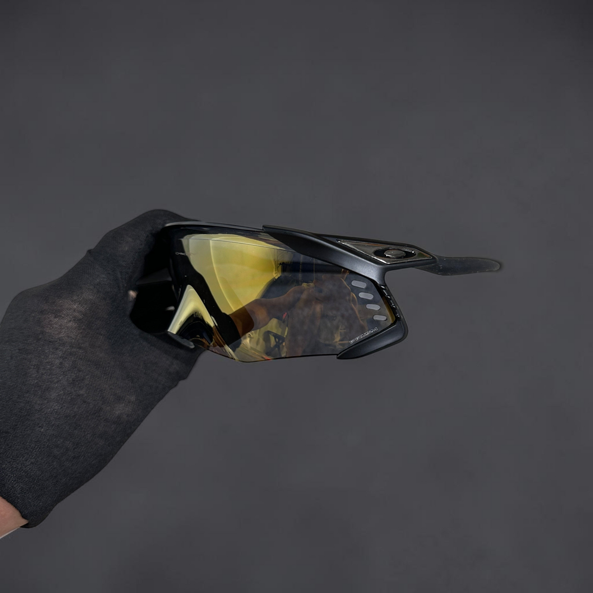 Gafas de sol deportivas OakGlasses Kato Velo con montura negra y lente de espejo dorada en mano con guante negro